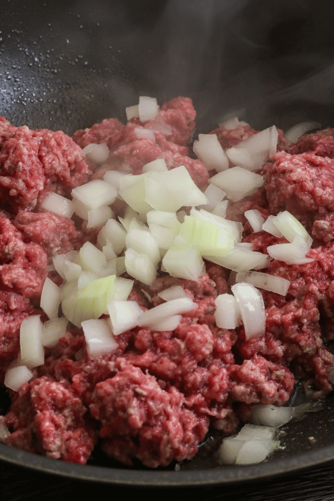 Raw ground beef and diced onions in a skillet beginning to cook for cowboy casserole.