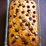 Moist pumpkin banana bread topped with chocolate chips in a clear glass loaf pan on a dark wooden surface.