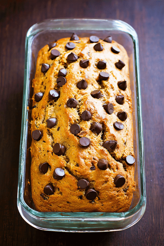 Moist pumpkin banana bread topped with chocolate chips in a clear glass loaf pan on a dark wooden surface.