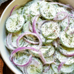 Creamy cucumber salad with red onions, dill, and black pepper in a white bowl on a rustic wooden surface.
