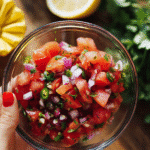Fresh tomato salsa in a glass bowl with diced tomatoes, onions, chilies, and herbs, held by hand over rustic background.