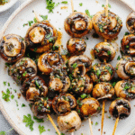 Grilled garlic mushroom skewers topped with herbs on a ceramic plate.