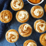 Freshly baked pumpkin cream cheese muffins with cream cheese swirls on a wire rack, styled with pumpkin puree and kitchen towel.