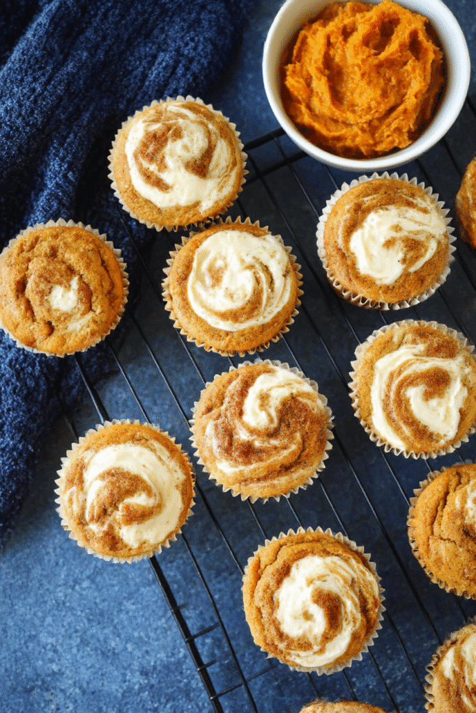 Freshly baked pumpkin cream cheese muffins with cream cheese swirls on a wire rack, styled with pumpkin puree and kitchen towel.