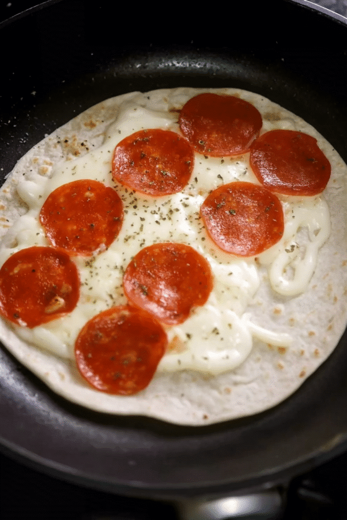 Flour tortilla in a skillet topped with melted cheese, pepperoni slices, and herbs, cooking for pepperoni pizzadillas.