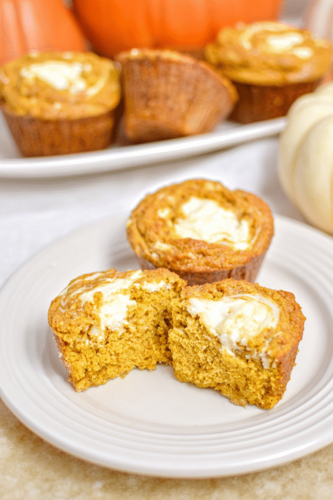 Halved keto pumpkin cream cheese muffin showing moist interior and creamy swirl, surrounded by whole muffins on a white plate.