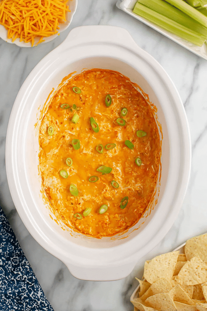 Crock Pot filled with finished Buffalo Chicken Dip topped with green onions, with celery, cheese, and chips on the side.