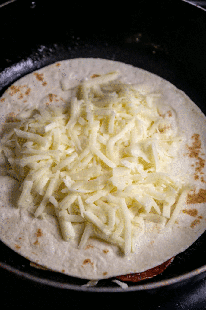 Shredded cheese on a flour tortilla in a skillet, ready to make pepperoni pizzadillas.