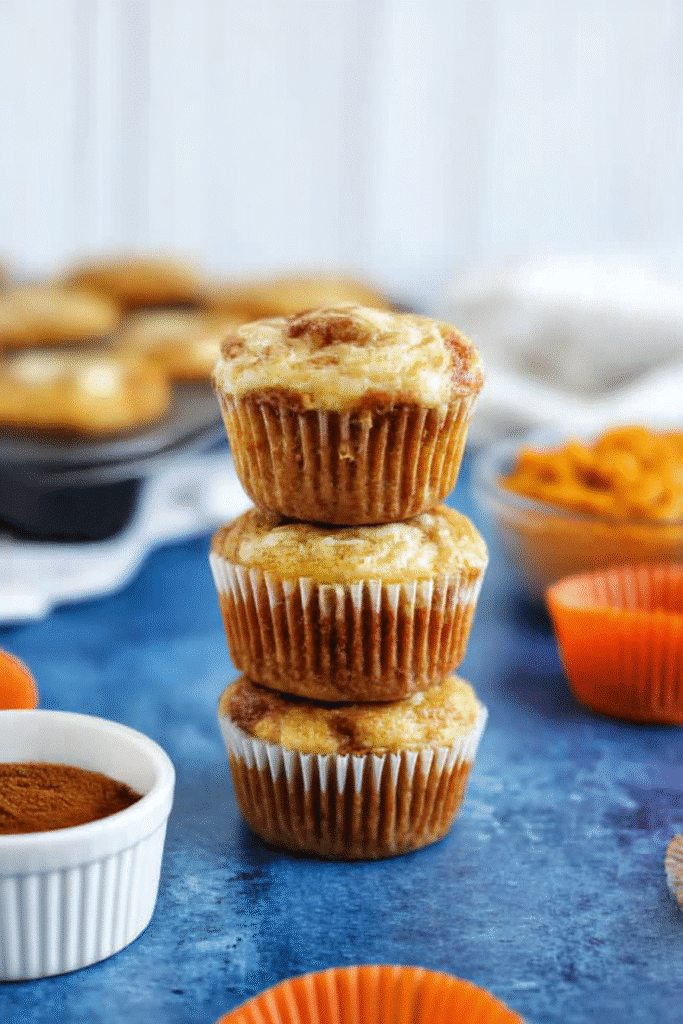 Stack of three pumpkin cream cheese muffins with visible swirls, surrounded by baking ingredients on a rustic blue surface.