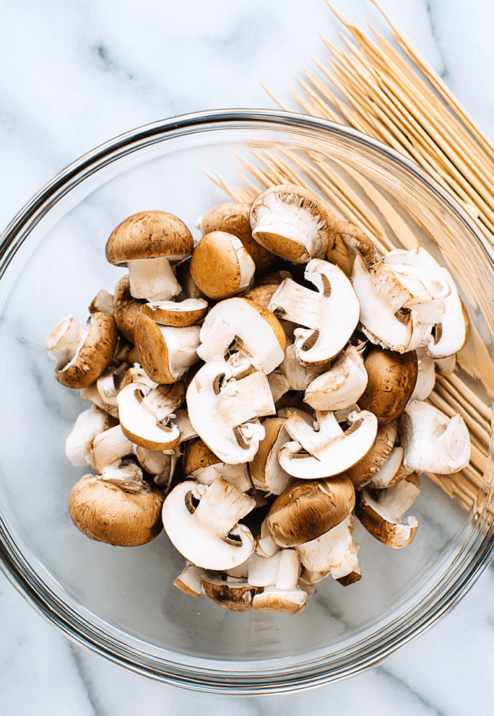 Halved cremini mushrooms in a bowl with skewers for grilled garlic mushroom skewers.