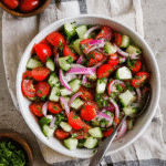 Fresh cucumber tomato salad in a white bowl with onions, parsley, and lemon on a beige navy-checkered cloth.