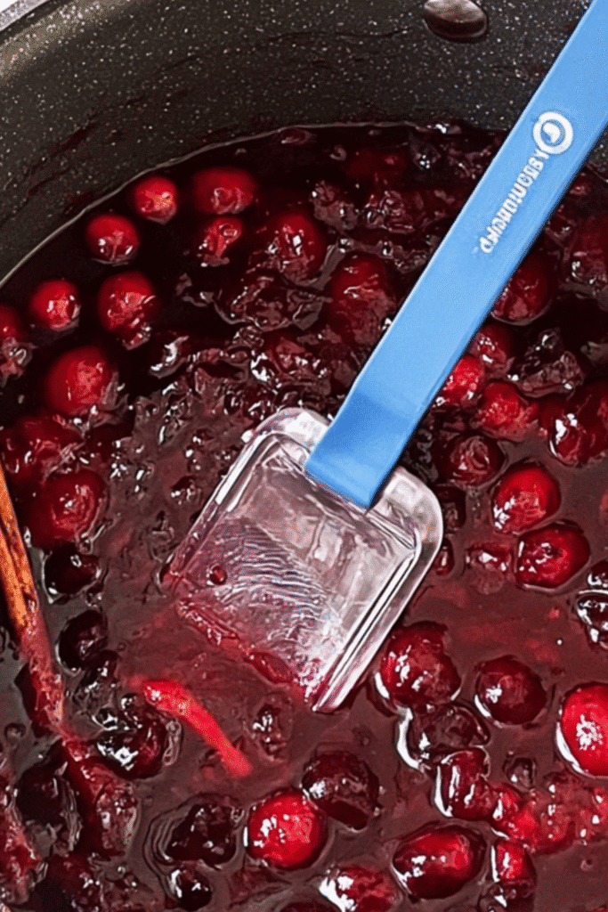 Cranberry jam cooking in a saucepan with a blue spatula and cinnamon stick