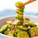 Spiral-cut Korean cucumber salad in a white bowl with chopsticks lifting a cucumber slice, topped with sesame seeds and chili seasoning.