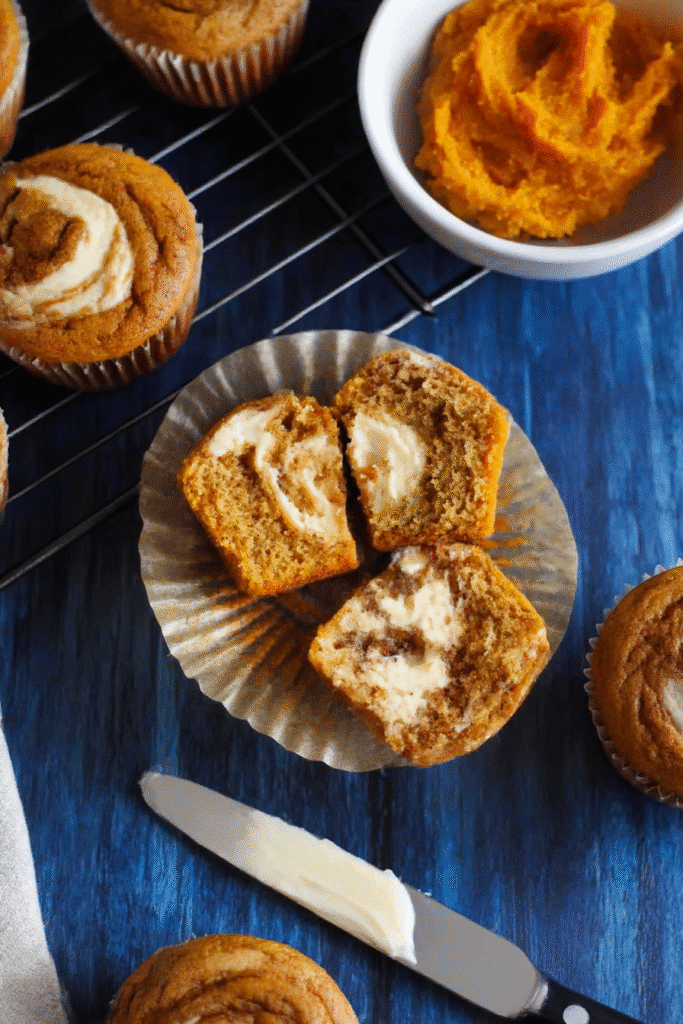 Halved pumpkin cream cheese muffin showing moist crumb and swirl detail, with a butter knife and pumpkin puree nearby.