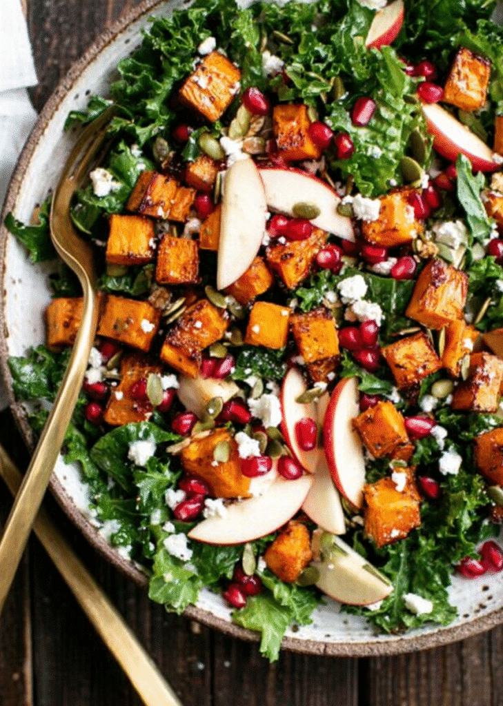 Mixed harvest salad with butternut squash, apple, kale, and pomegranate, served with golden utensils.