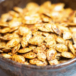 Golden-brown roasted pumpkin seeds in a rustic ceramic bowl, seasoned with spices for a crunchy autumn snack.
