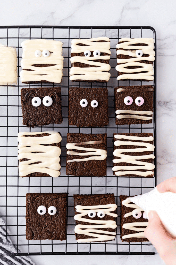 A hand pipes zigzag white frosting onto chocolate brownies with candy eyes, transforming them into adorable Halloween mummy brownies.