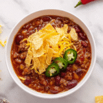 Easy Crockpot Chili topped with cheese, tortilla chips, and jalapeño slices in a white bowl.