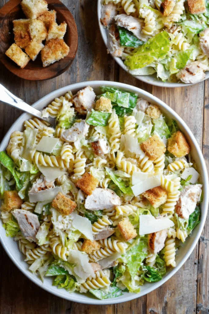 Chicken Caesar Pasta Salad with rotini, lettuce, chicken, croutons, and parmesan in a white bowl on a rustic wood table.