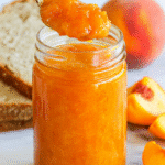 Fresh peach jam in a glass jar with spoon, rustic bread, and ripe peaches on marble.