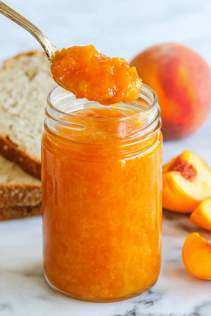 Fresh peach jam in a glass jar with spoon, rustic bread, and ripe peaches on marble.