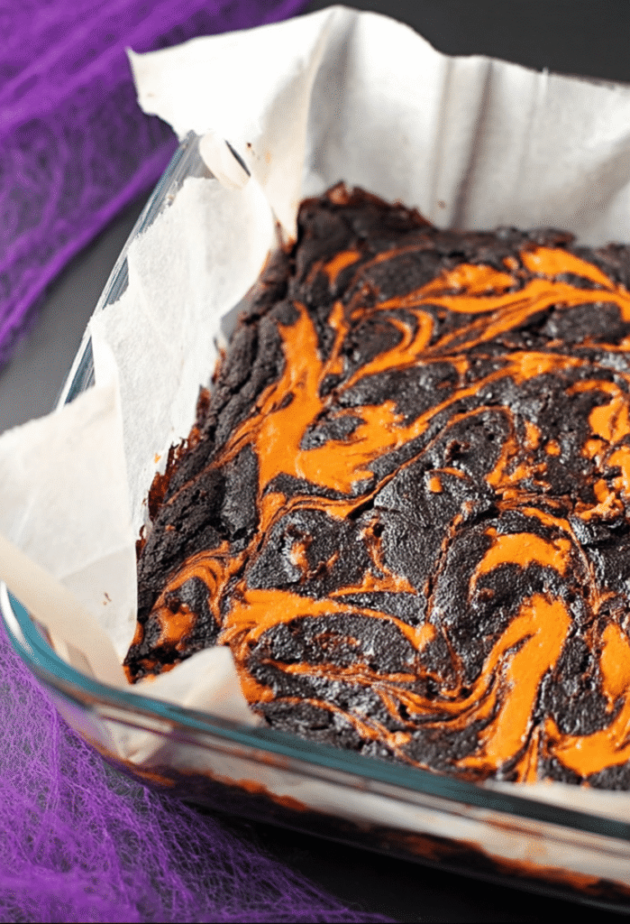 Glass dish of Halloween swirl cream cheese brownies with vivid orange and black marbled top, lined with parchment paper.