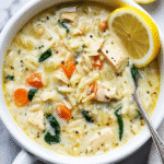 Bowl of creamy lemon chicken orzo soup with spinach, carrots, orzo, and a lemon wedge.