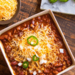 Bowl of slow cooker chili recipe topped with shredded cheddar, onions, and jalapeños on rustic wood background.