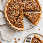Overhead shot of a praline pumpkin pie with a slice removed, topped with crunchy pecans and set on a light background.