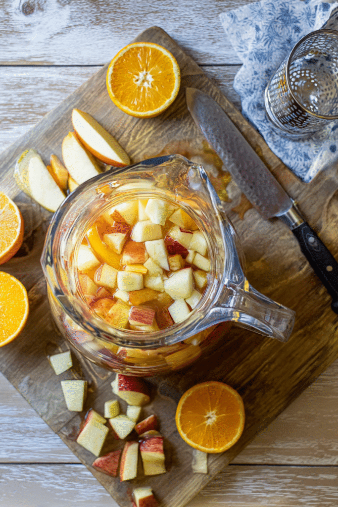 Overhead view of apple cider sangria preparation with apples and oranges on a rustic cutting board.
