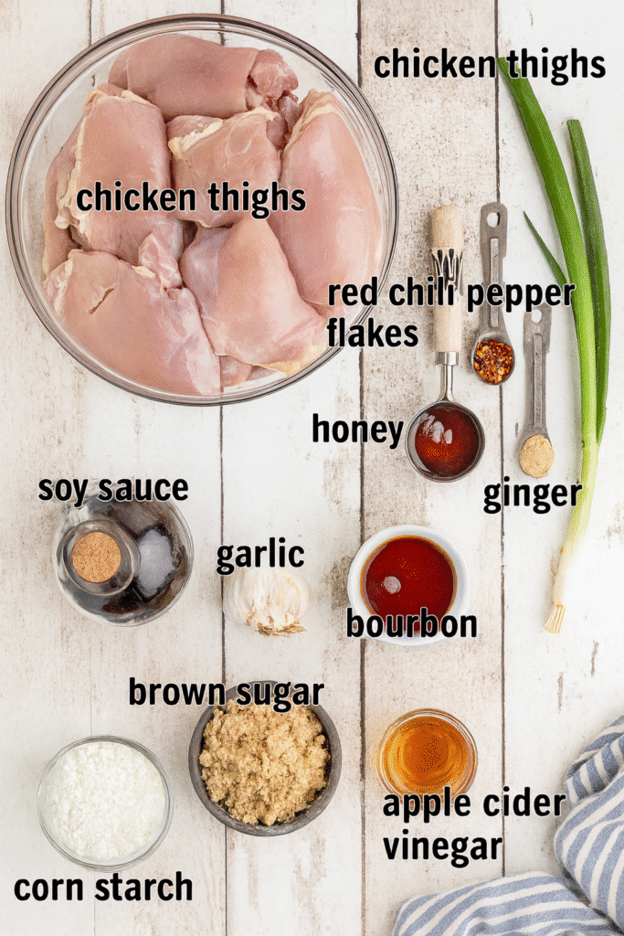 Flat lay of crock pot bourbon chicken ingredients including chicken thighs, bourbon, garlic, soy sauce, and spices on a rustic white surface.