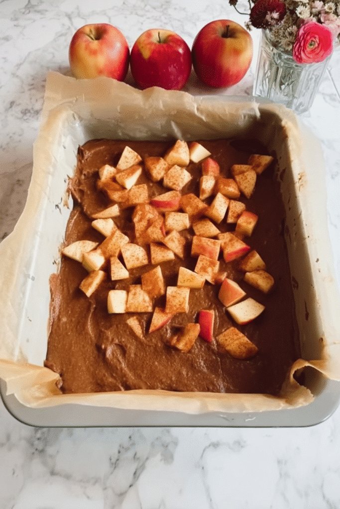 Cinnamon-coated apples layered over brown butter batter in a parchment-lined baking dish.