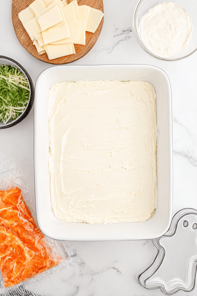 Layer of cream cheese spread in baking dish for Halloween Buffalo Chicken Dip, surrounded by festive ingredients.