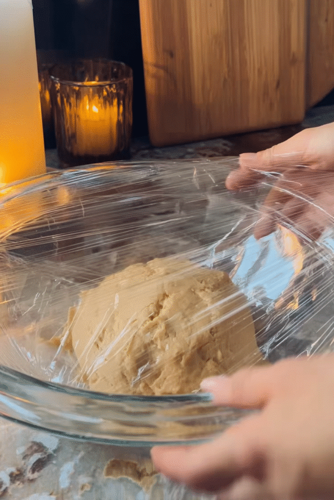 Apple Pie Cheesecake Brioche dough in a glass bowl being covered with plastic wrap, ready to rise on a cozy kitchen counter.