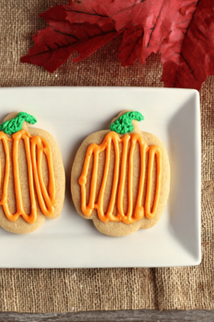 Two pumpkin spice sugar cookies with orange frosting on a white plate with fall decor. Title: Pumpkin Spice Sugar Cookies Styled for Fall