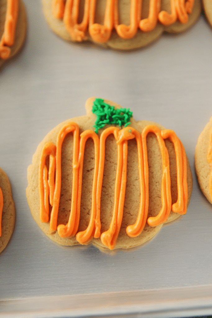 Pumpkin spice sugar cookie shaped like a pumpkin, topped with vibrant orange frosting and a green stem, placed on a metal baking sheet.