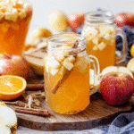 Apple cider sangria in mason jars with apple chunks and cinnamon, styled with fresh fruit on a rustic wooden board.