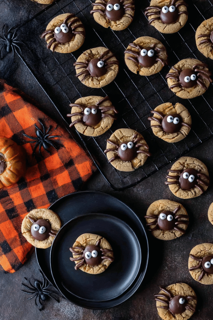 Peanut butter spider cookies styled on black plates and a wire rack with chocolate legs, candy eyes, and Halloween decor on a rustic dark background.