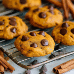 Pumpkin chocolate chip cookies cooling on a wire rack with cinnamon sticks and scattered chocolate chips.