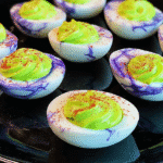 Halloween deviled eggs with purple marble-veined egg whites, neon green filling, and paprika garnish on a black platter.