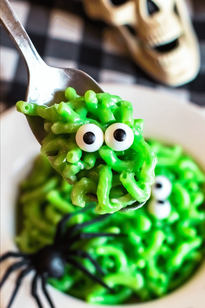 Spoonful of monster mac and cheese with candy eyeballs, Halloween-themed background with skeleton and spider.