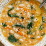 A bowl of the best white bean soup with spinach, carrots, and creamy broth on a rustic wooden table