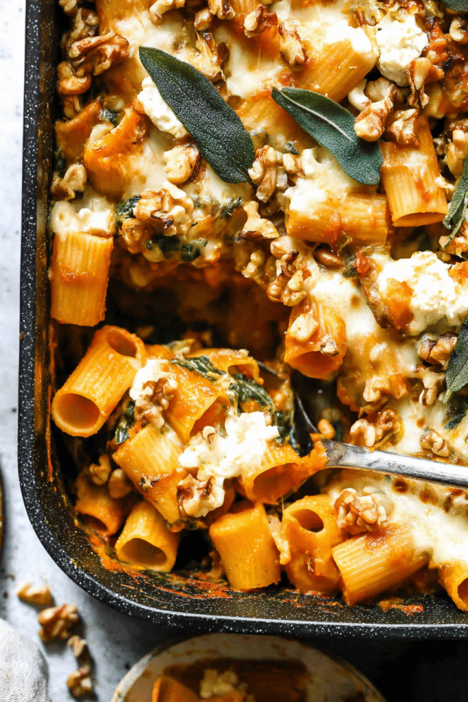 Cheesy pumpkin pasta bake with rigatoni, walnuts, and sage being scooped from the pan.