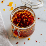 Glass jar of homemade chili oil with red pepper flakes on a white countertop.