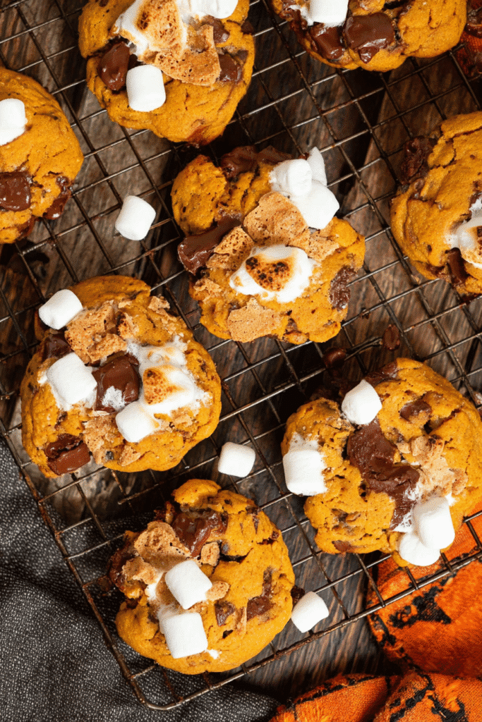 Overhead view of pumpkin s’mores cookies with marshmallows, chocolate chips, and graham cracker cooling on a wire rack.