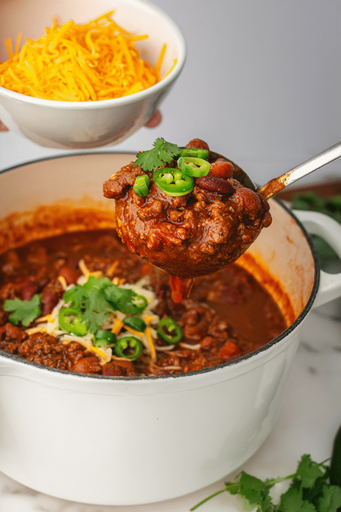 Ladle serving homemade stovetop chili with cheese and cilantro into a white bowl.