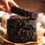 Hand lifting a slice of moist chocolate pumpkin bread stacked with melty chocolate chips.