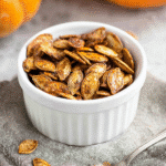 Pumpkin seeds recipe with roasted golden seeds in a white bowl and spoon on rustic cloth with pumpkin background.