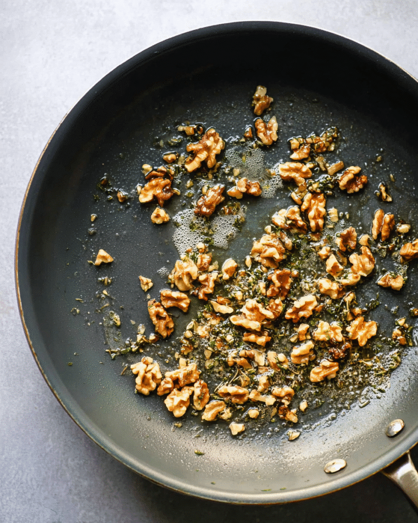Walnuts toasted with butter and herbs in a skillet for pumpkin pasta bake.