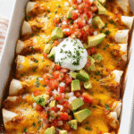 Cheesy beef enchiladas topped with avocado, salsa, and sour cream in a white baking dish.